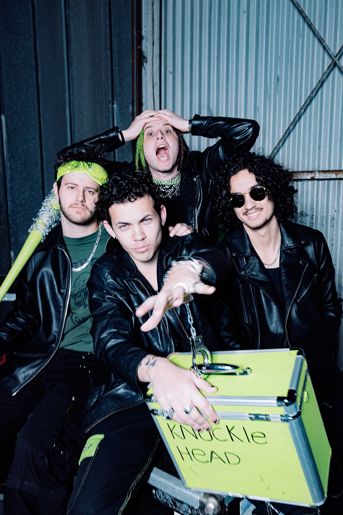 Four people posing with a suitcase labeled 'Knuckle Head' against a metallic wall.